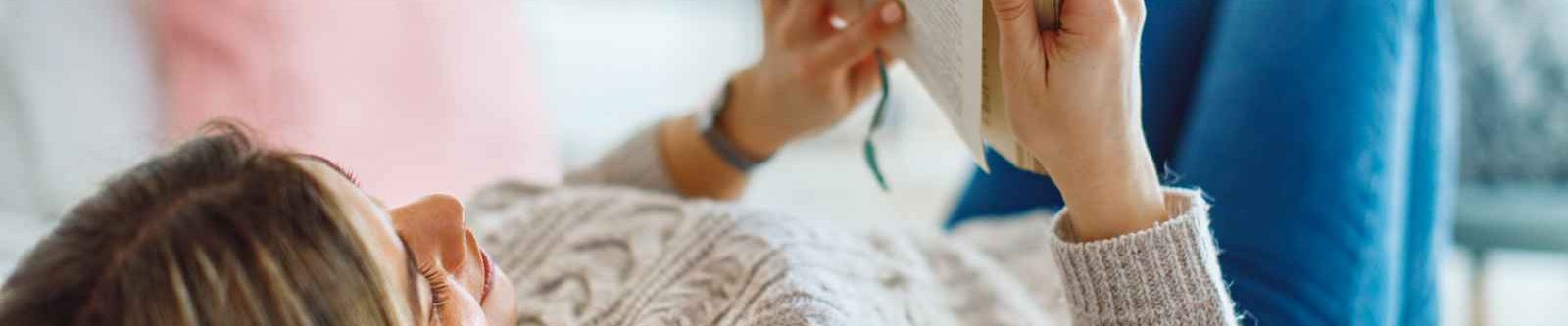 Young woman lies on her back in bed reading a book