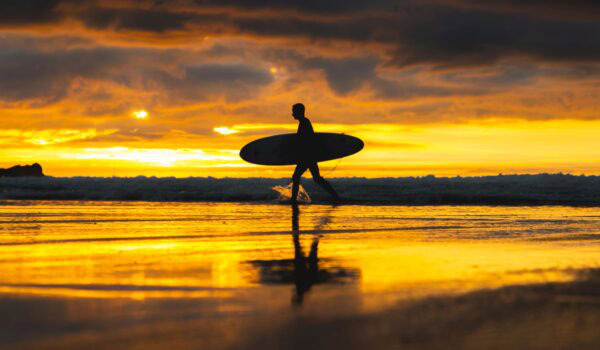 Lone surfer walking across Fistral Beach at sunset
